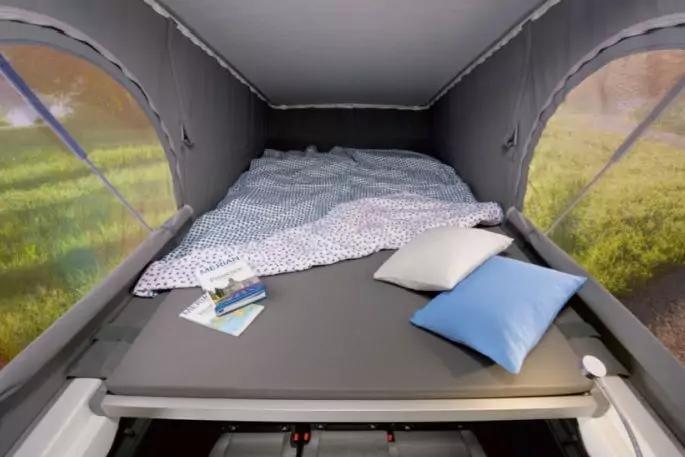 Innenraum eines Dachzelts mit Matratze, gemustertem Bettbezug, zwei Kissen (blau, wei&szlig;), zwei Brosch&uuml;ren auf der Matratze, seitliche Fenster mit gr&uuml;ner Wiese sichtbar, graue Innenw&auml;nde und Decke