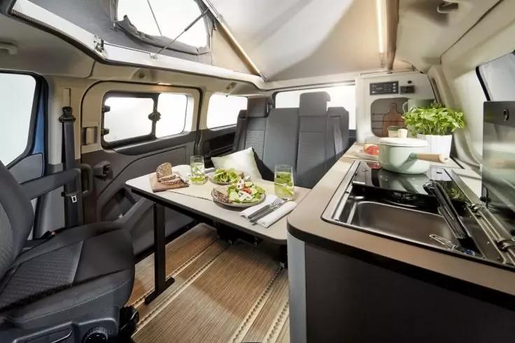 Innenraum eines Campers mit Esstisch f&uuml;r zwei, Sitzb&auml;nken, K&uuml;chenzeile mit Sp&uuml;le und Kochfeld, Schalen mit Salat auf dem Tisch, Fenster und Dachluke