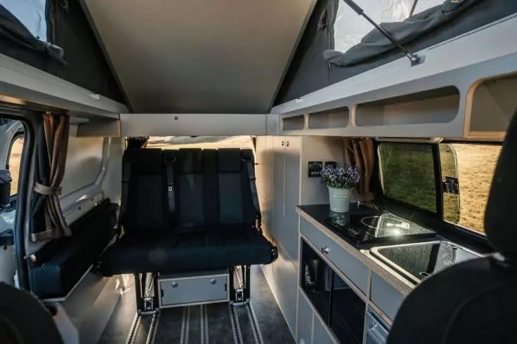 Innenraum eines Camping-Vans mit zweisitziger Bank, K&uuml;chenzeile mit Sp&uuml;le und Herd, Fenster mit Vorhang, kleine Topfpflanze, Staur&auml;ume und aufgeklapptes Aufstelldach