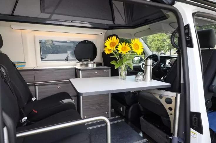 Innenraum eines Campingbusses mit ge&ouml;ffneter Seitent&uuml;r; Tisch mit Vase mit Sonnenblumen und Thermoskanne, gedrehte Vordersitze, K&uuml;chenzeile mit Sp&uuml;le, Schr&auml;nken und Fenster