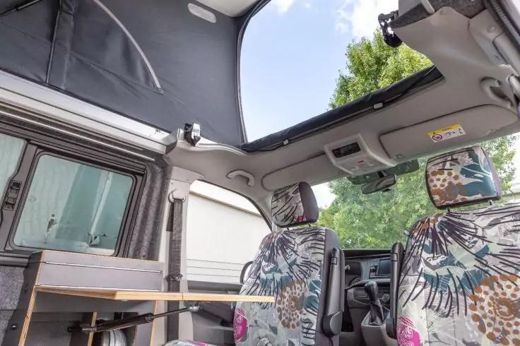 Innenraum eines Vans mit ge&ouml;ffnetem Aufstelldach, zwei vordere Sitze mit gemusterten Bez&uuml;gen, kleiner Seitentisch, Fenster mit B&auml;umen drau&szlig;en, Himmel durch Dach&ouml;ffnung sichtbar