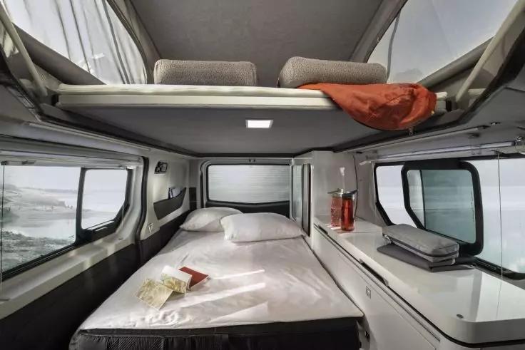 Innenraum eines Wohnmobils, Schlafbereich mit unterem Bett mit wei&szlig;er Bettw&auml;sche und Kissen, oberer Schlafkoje mit orangefarbener Decke, seitliche Fenster mit Blick auf Wasser und Ufer, Ablage mit zwei roten Trinkflaschen und geschlossenem Laptop, ve