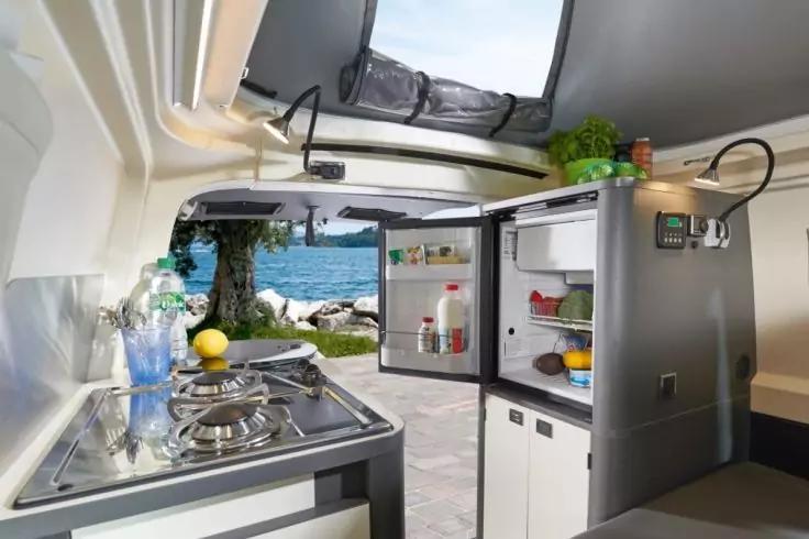 Innenansicht eines Wohnmobils: K&uuml;chenzeile mit Gaskocher, Sp&uuml;le, ge&ouml;ffnetem K&uuml;hlschrank mit Lebensmitteln und Flaschen; offene Heckklappe mit Blick auf Meer, felsiges Ufer und Baum; Hochbett und Kr&auml;utertopf sichtbar