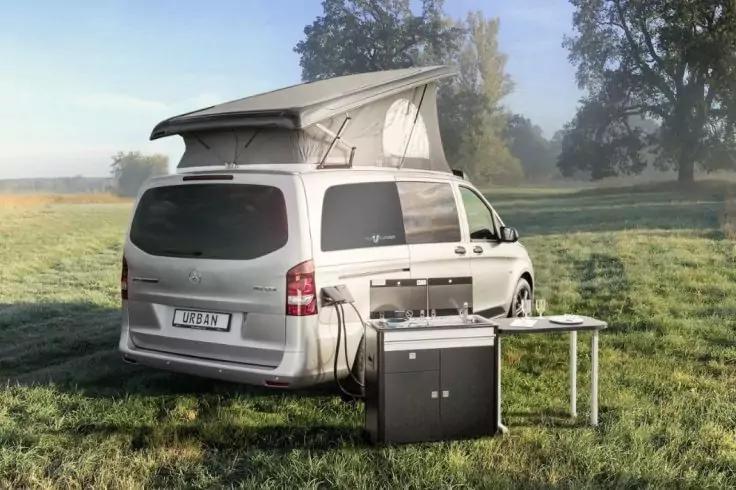 Silberner Camper mit aufgestelltem Aufstelldach auf einer Wiese, mobile K&uuml;cheneinheit und kleiner Tisch daneben, B&auml;ume im Hintergrund