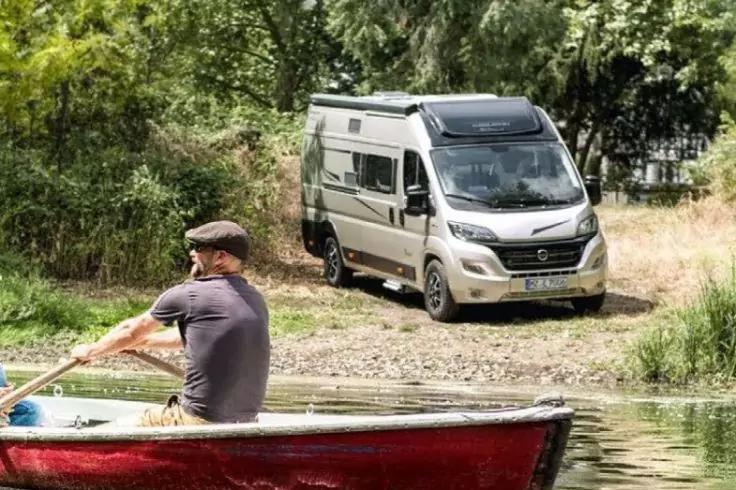 Person in einem Ruderboot paddelt auf dem Wasser; im Hintergrund Wohnmobil auf kiesigem Ufer vor B&auml;umen