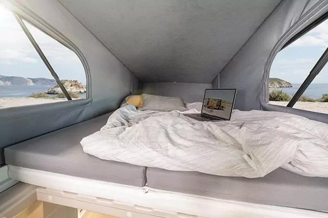 Schlafbereich mit ungemachtem Bett, wei&szlig;er Decke, Kissen und offenem Laptop; zwei Fenster zeigen Meer und K&uuml;ste