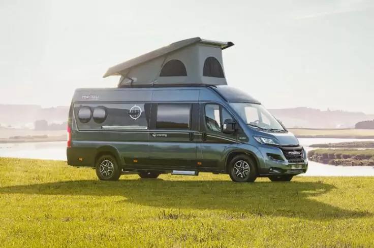 Dunkelgr&uuml;ner Campingbus mit ge&ouml;ffnetem Aufstelldach, geparkt auf einer Wiese neben einem Gew&auml;sser; flaches Ufer und Himmel im Hintergrund