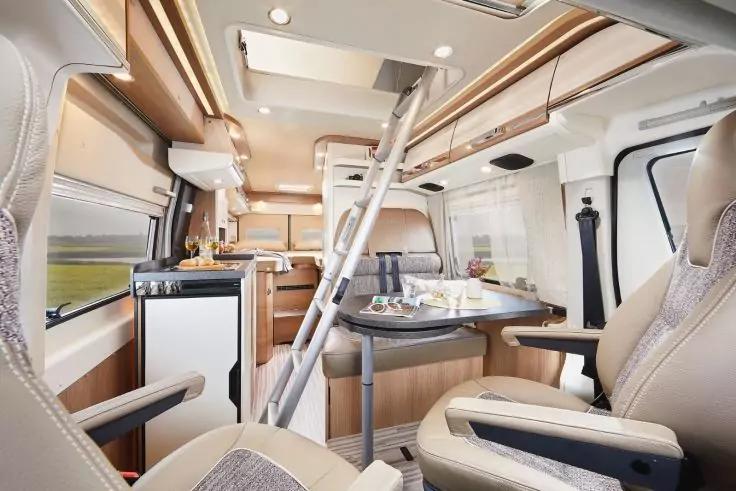 Innenraum eines Wohnmobils mit zwei drehbaren Vordersitzen, Esstisch mit Tellern und Gl&auml;sern, Leiter zur oberen Schlafkoje, K&uuml;chenzeile mit Sp&uuml;le, Herd und K&uuml;hlschrank, Fenstern und Deckenleuchten