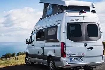 Silberner Camper mit aufgeklapptem Aufstelldach, geparkt an einer K&uuml;stenstra&szlig;e; Meer, Gras und bew&ouml;lkter Himmel im Hintergrund