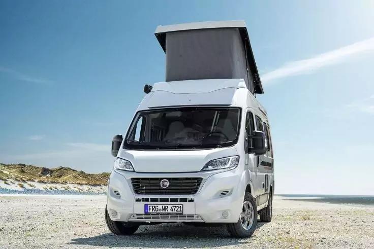 Wei&szlig;er Camper mit aufgeklapptem Aufstelldach, Frontal‑Schr&auml;gaufnahme, auf sandigem Strand mit D&uuml;nen und Meer im Hintergrund