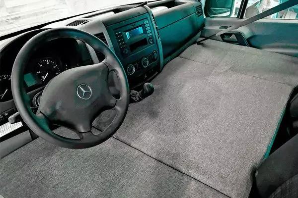 Innenraum eines Fahrzeugs: Lenkrad mit Mercedes-Logo, Armaturentafel mit Display und Bedienelementen, Schalthebel, graue Polsterfl&auml;che &uuml;ber Mittelkonsole und Beifahrersitz, Vordersitze und T&uuml;rverkleidung