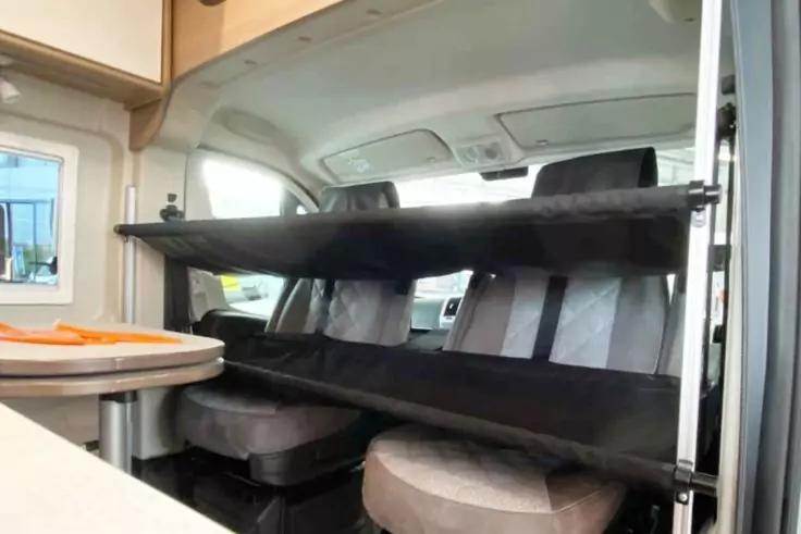 Innenraum eines Wohnmobils mit zwei Vordersitzen, dar&uuml;ber zwei schwarze Stoffplattformen auf einem Metallrahmen, runder Tisch links, Oberschr&auml;nke und Fenster