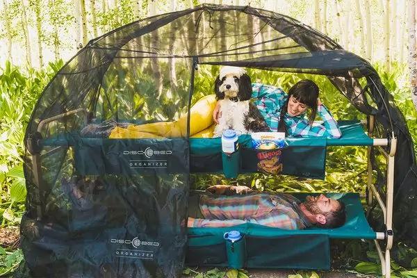 Zweist&ouml;ckiges Feldbett mit schwarzem Moskitonetz, zwei Kinder, wei&szlig;-brauner Hund sitzt oben, Snackdose und Becher, Holzzaun und Pflanzen im Hintergrund