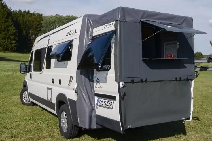 Wei&szlig;er Campervan auf einer Wiese, grauer Stoff‑Heckaufbau mit ge&ouml;ffneten Klappen, seitliche Fenster ge&ouml;ffnet, B&auml;ume im Hintergrund