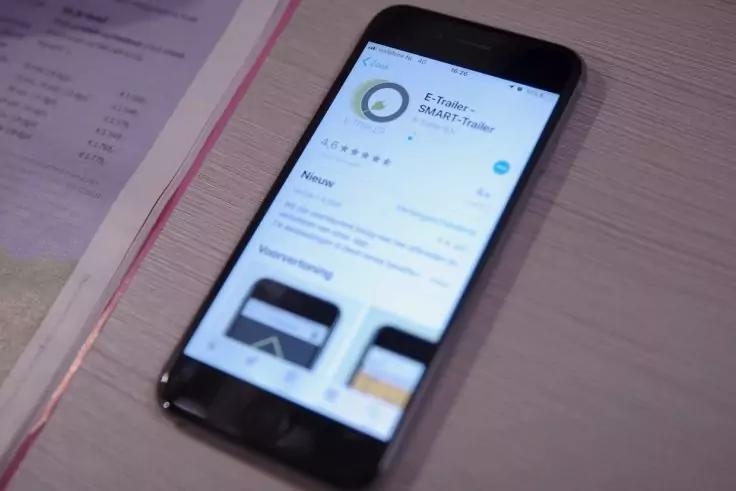 Smartphone liegt auf einem Tisch; auf dem Bildschirm ist eine App-Detailseite mit App-Symbol, Titel, Text und Vorschaubildern sichtbar; neben dem Telefon liegt ein Blatt Papier