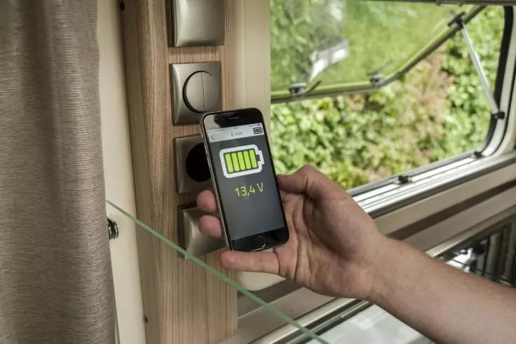Hand hält Smartphone mit grünem Batterie-Symbol und "13.4V" vor einer Wand mit Steckdose, neben offenem Fenster mit grüner Vegetation und Gardine