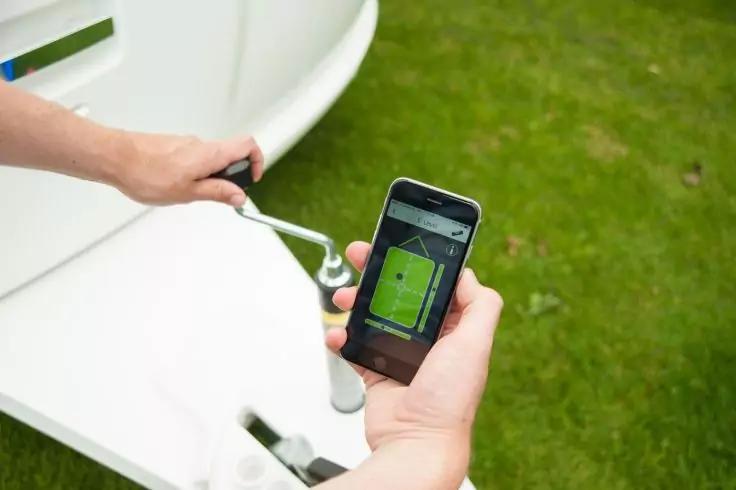 Eine Hand hält ein Smartphone, auf dessen Display eine grüne Fläche mit einer kleinen Flagge zu sehen ist; die andere Hand dreht eine Metallkurbel an einem weißen Gerät; grüner Rasen im Hintergrund