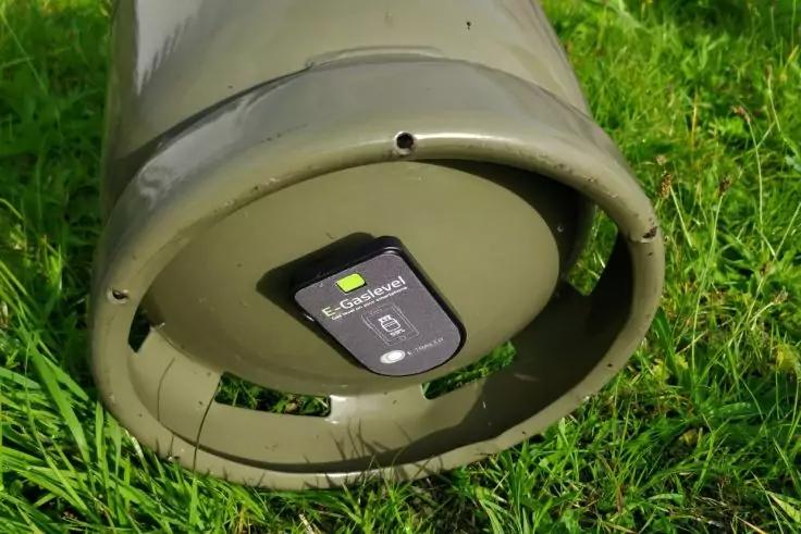 Grüner Metallzylinder liegt auf Gras; am unteren Ende mittig ein schwarzes Gerät mit sichtbarem Etikett befestigt