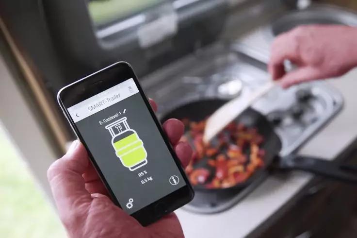 Vordergrund: Hand hält Smartphone mit angezeigtem Flaschen-/Gasflaschen-Symbol und Prozentanzeige; im Hintergrund rührt eine andere Hand mit einem Holzspatel Gemüse in einer Pfanne auf einem Herd