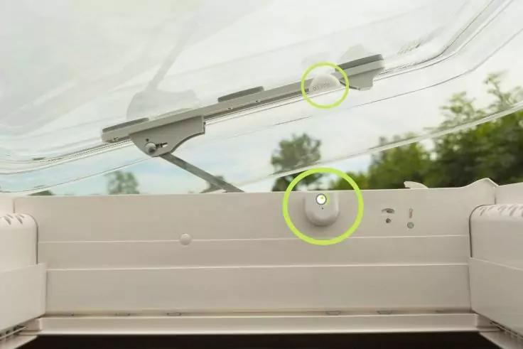 Innenansicht eines geöffneten Dachfensters mit sichtbarem Scharnier, Metallgestänge und Dichtung; zwei grüne Kreise markieren Stellen; Bäume und bewölkter Himmel durchs Glas sichtbar