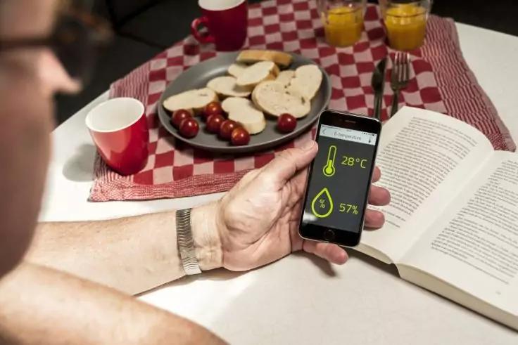 Person hält Smartphone mit Anzeige 28°C und 57% neben offenem Buch; Teller mit Brotscheiben und Kirschtomaten, zwei Gläser Orangensaft, rote Becher und rot-weiß kariertes Tuch auf dem Tisch