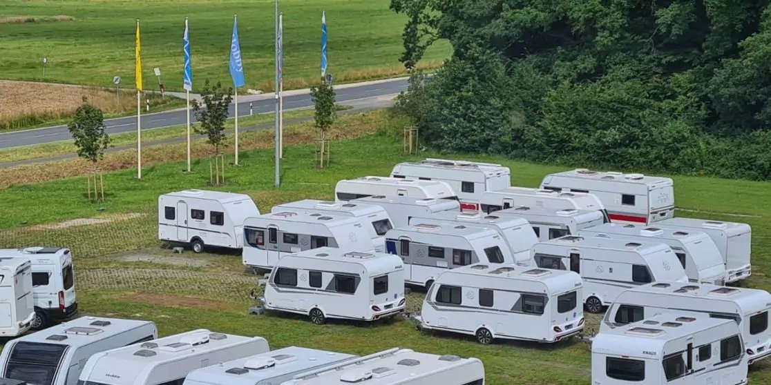 Mehrere wei&szlig;e Wohnwagen dicht auf einer Wiese, im Hintergrund B&auml;ume, eine Stra&szlig;e und mehrere Flaggen