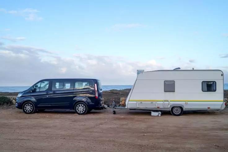 Schwarzer Van mit angehängtem weißem Caravan auf unbefestigtem Boden, Meer und Himmel im Hintergrund