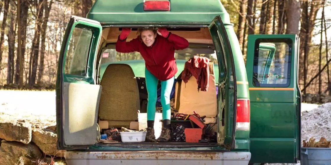 Offener gr&uuml;ner Van von hinten; eine Person in rotem Pullover und t&uuml;rkisfarbenen Leggings lehnt aus dem Laderaum; im Innenraum h&auml;ngen Kleidungsst&uuml;cke, auf dem Boden liegen Kisten und Beh&auml;lter; B&auml;ume im Hintergrund