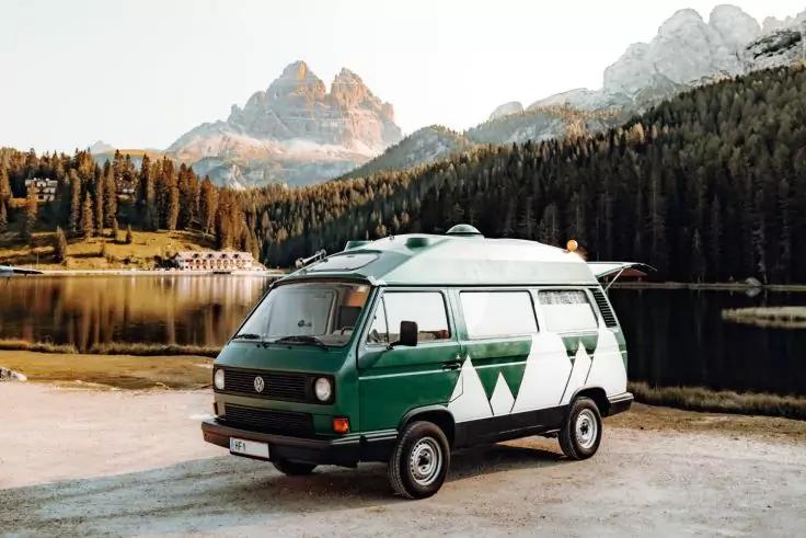 Gr&uuml;ner Camper-Van mit wei&szlig;em Dreiecksmuster, am Ufer eines Bergsees geparkt, im Hintergrund Nadelwald und Berge