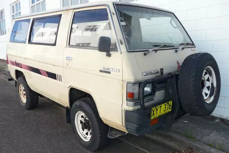 Beiger Kleinbus mit vorne montiertem Ersatzrad und schwarzem Seitenstreifen, geparkt neben einer wei&szlig;en Fassade