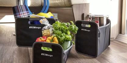 Drei schwarze Stoff-Aufbewahrungsboxen auf Holzboden neben Sofa und Fenster; eine enth&auml;lt einen Topf mit gr&uuml;ner Zimmerpflanze und Gem&uuml;se (Paprika, Tomaten), eine ein gefaltetes Plaid und ein gelbes Objekt, die dritte einen Metalltopf