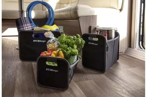 Drei schwarze Aufbewahrungsboxen mit wei&szlig;em Berger-Logo auf Holzboden neben einer hellen Polstersitzbank und einem Fenster; links kariertes Tuch und blauer Schlauch, Mitte Topfpflanze mit gr&uuml;nen Bl&auml;ttern sowie rote und gelbe Paprika und Zucchini, rec