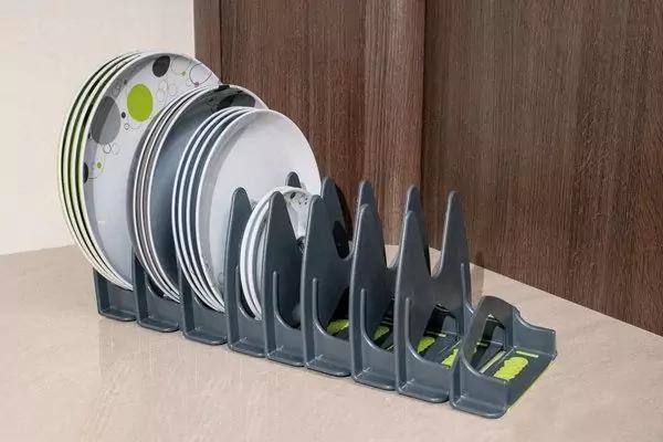 Grauer Kunststoff-Tellerst&auml;nder auf Arbeitsfl&auml;che vor dunklem Holzpaneel, mehrere Teller unterschiedlicher Gr&ouml;&szlig;e (wei&szlig;e Teller mit gr&uuml;nem Muster, wei&szlig;e und graue Teller) stehend