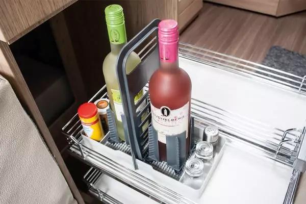 Zwei Glasflaschen (eine gr&uuml;n, eine matt mit rosa Verschluss) in einem grauen Trageeinsatz auf einem ausziehbaren Metallgitterkorb; daneben ein gelbes D&ouml;schen und zwei kleine Gl&auml;ser