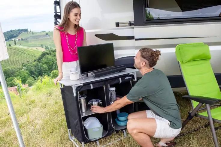 Frau steht neben einer mobilen Campingk&uuml;che mit Flachbildschirm auf der Arbeitsfl&auml;che; Mann kniet davor und greift in ein offenes Fach; Wohnmobil, gr&uuml;ner Klappsessel und h&uuml;gelige Wiese im Hintergrund