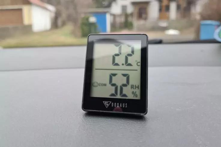 Schwarzes digitales Thermometer/Hygrometer auf dem Armaturenbrett eines Autos, Display zeigt "2.2&deg;C" oben und "52 %" unten; unscharfer Hintergrund mit H&auml;usern
