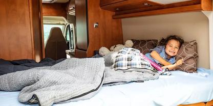 Person liegt l&auml;chelnd auf einem Bett im Wohnmobil, mit Decken, Kissen und einem Stofftier; Holzinnenraum und Fahrerhaus im Hintergrund