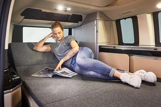 Person liegt auf umgeklapptem Sitz/Schlaffläche in einem Van, gestützt auf einen Ellbogen, liest ein geöffnetes Magazin; trägt T‑Shirt, Jeans und weiße Sneaker; Van-Innenraum mit Fenstern und Deckenlampen