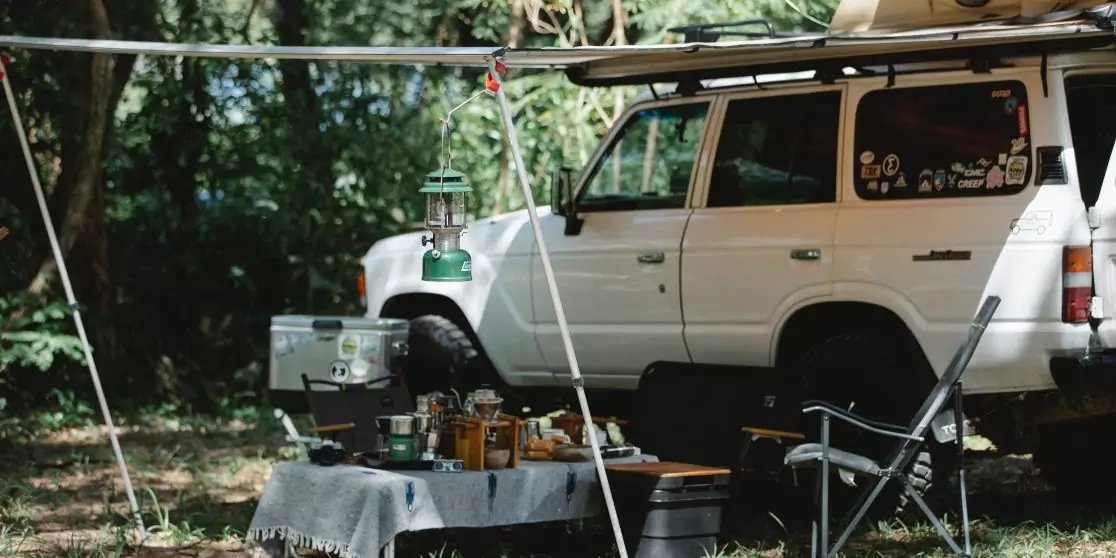 Wei&szlig;er Gel&auml;ndewagen neben einem Tisch mit Flaschen, Laterne, Gaskocher und Klappstuhl unter B&auml;umen