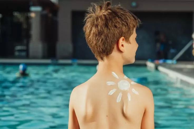 Junge ohne Oberteil von hinten, mit kurzen braunen Haaren und Sonnenschutzcreme in Form einer Sonne auf Schulter und oberem Rücken, steht am Rand eines Schwimmbeckens; im Hintergrund Wasser und unscharfe Schwimmer