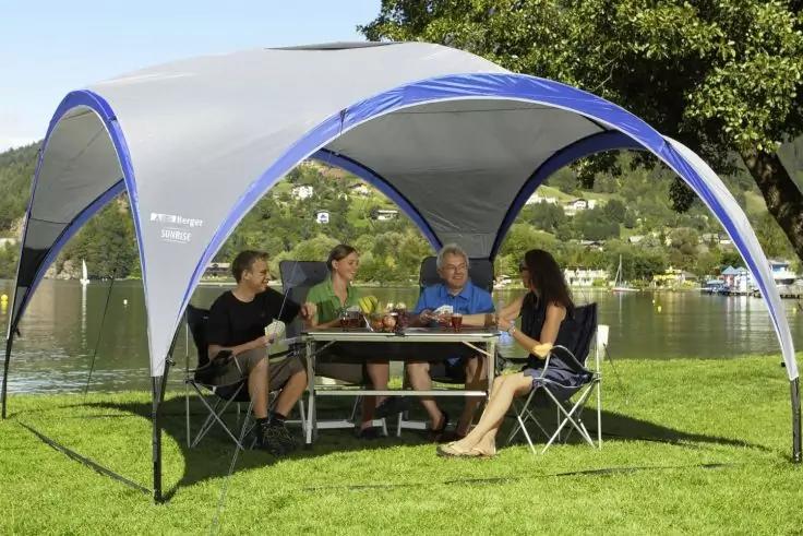 Vier Personen sitzen an einem Picknicktisch unter einem blau-weißen Pavillon auf einer Wiese; im Hintergrund See, Boote und Bäume