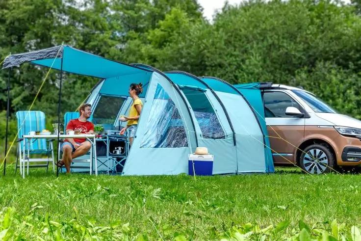 Auto mit angebautem Vorzelt; zwei Personen an Campingtisch, Campingstühle, Kühlbox, Wiese und Bäume
