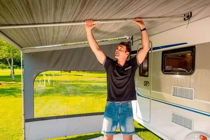 Person h&auml;lt mit beiden H&auml;nden die Stange einer Markise an einem Wohnwagen, tr&auml;gt schwarzes Polo und Jeansshorts, Rasen und B&auml;ume im Hintergrund