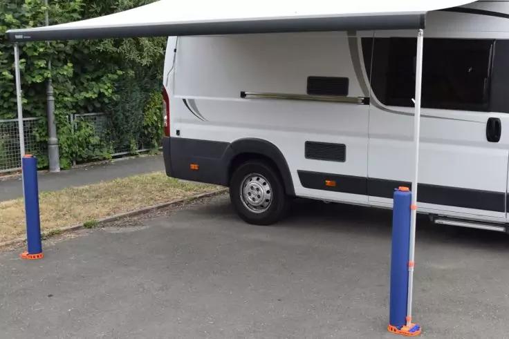 Wei&szlig;es Wohnmobil seitlich mit ausgefahrener Markise, zwei blaue St&uuml;tzen, hinteres Rad sichtbar, geparkt auf Asphalt neben Gras, Zaun und B&auml;umen im Hintergrund