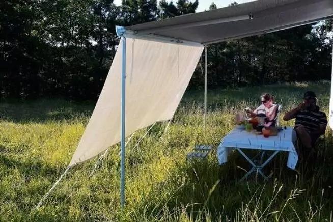 Aufgespannte Plane als Sonnendach auf Metallstangen &uuml;ber einem Tisch mit wei&szlig;er Tischdecke in hohem Gras; zwei Personen sitzen am Tisch; B&auml;ume im Hintergrund