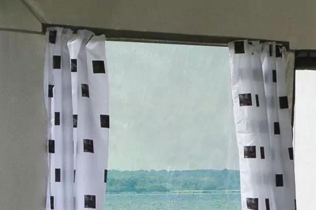 Fensterrahmen mit wei&szlig;en Gardinen mit schwarzen Quadraten; im Fenster Meer und Himmel am Horizont