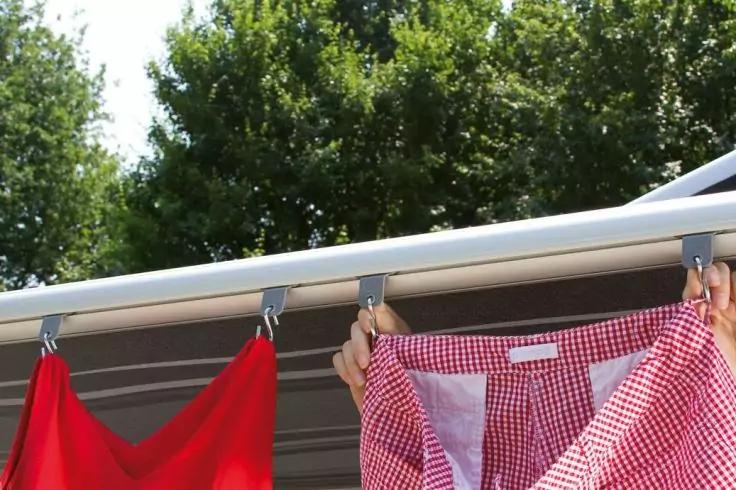 Rot-wei&szlig; karierte Bluse an W&auml;scheleine, oben von zwei H&auml;nden gehalten; links rotes Tuch, Metallhaken, Markisenleiste, B&auml;ume im Hintergrund