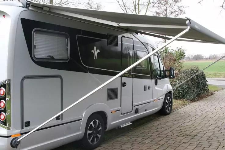 Wei&szlig;-graues Wohnmobil mit ausgefahrener Markise, geparkt auf gepflastertem Untergrund; B&uuml;sche rechts, Feld und B&auml;ume im Hintergrund
