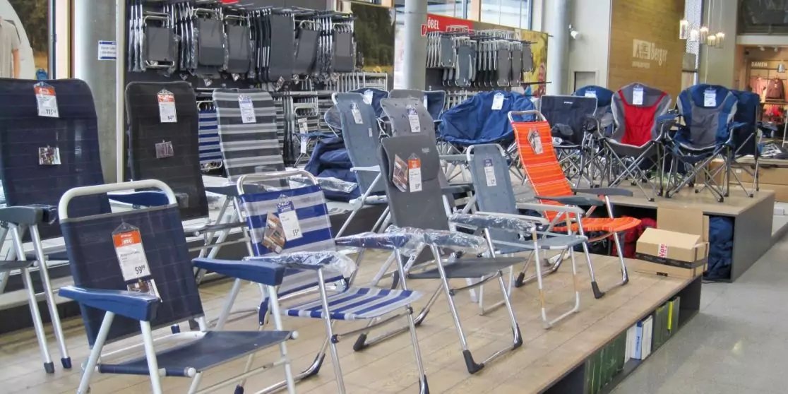 Reihe zusammenklappbarer Campingst&uuml;hle in verschiedenen Farben (blau, schwarz, orange) auf einer Verkaufsfl&auml;che im Laden mit Preisschildern