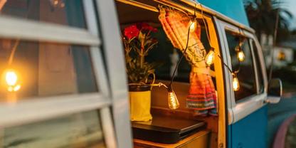 Geparkter blauer Campingbus mit ge&ouml;ffneter Schiebet&uuml;r; innen ein Tisch mit Topfrose, ein gemusteter Vorhang und Lichterkette
